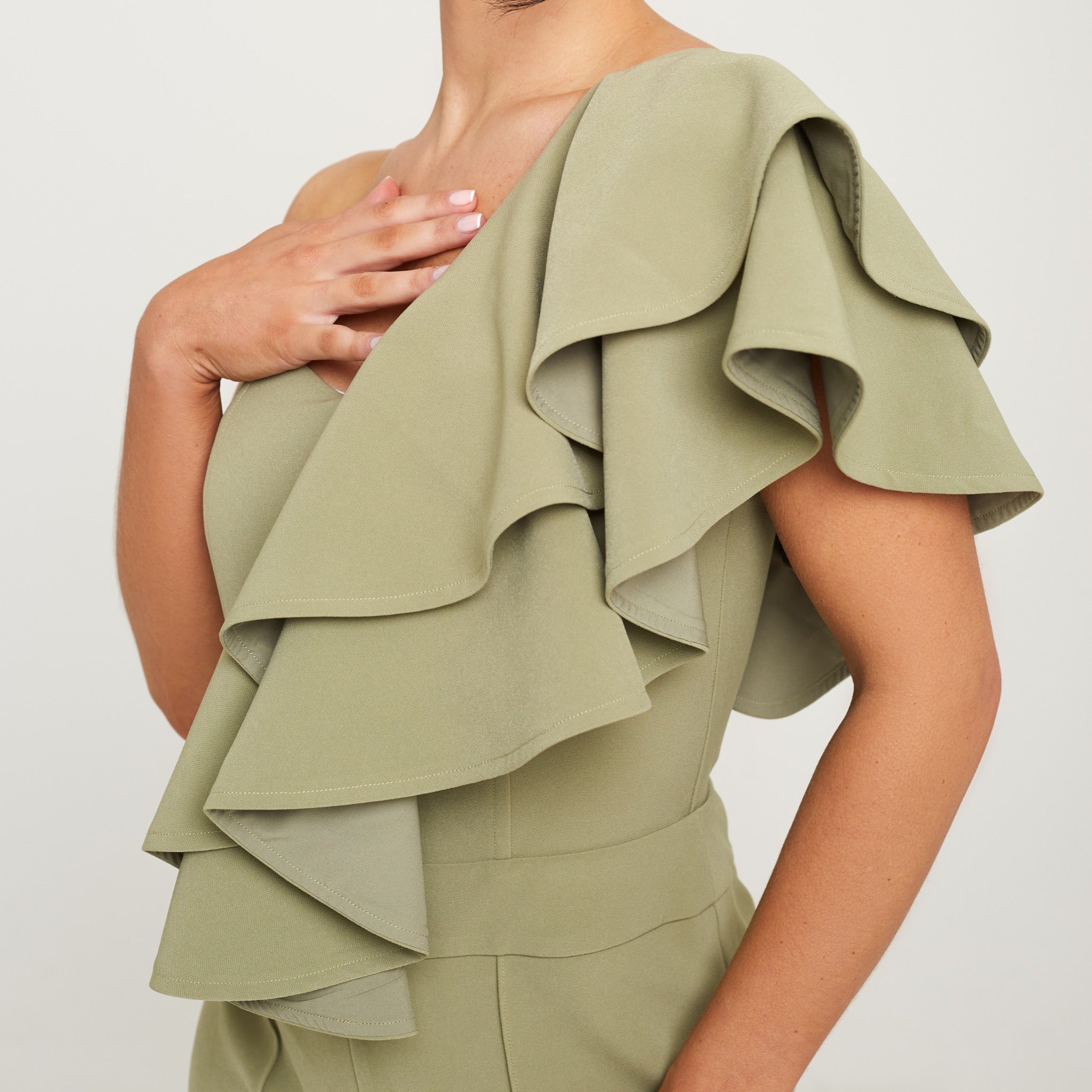 Cally Jane Beech, a woman with shoulder-length brunette hair, showcases a stylish sage green one-shoulder ruffle jumpsuit with cigarette trousers. The jumpsuit features a single shoulder strap, creating an asymmetrical neckline, while the ruffle detail cascades elegantly down the bodice, adding a feminine and playful touch. The jumpsuit's sleek cigarette trousers fit snugly and taper down to the ankles, creating a streamlined and polished look.