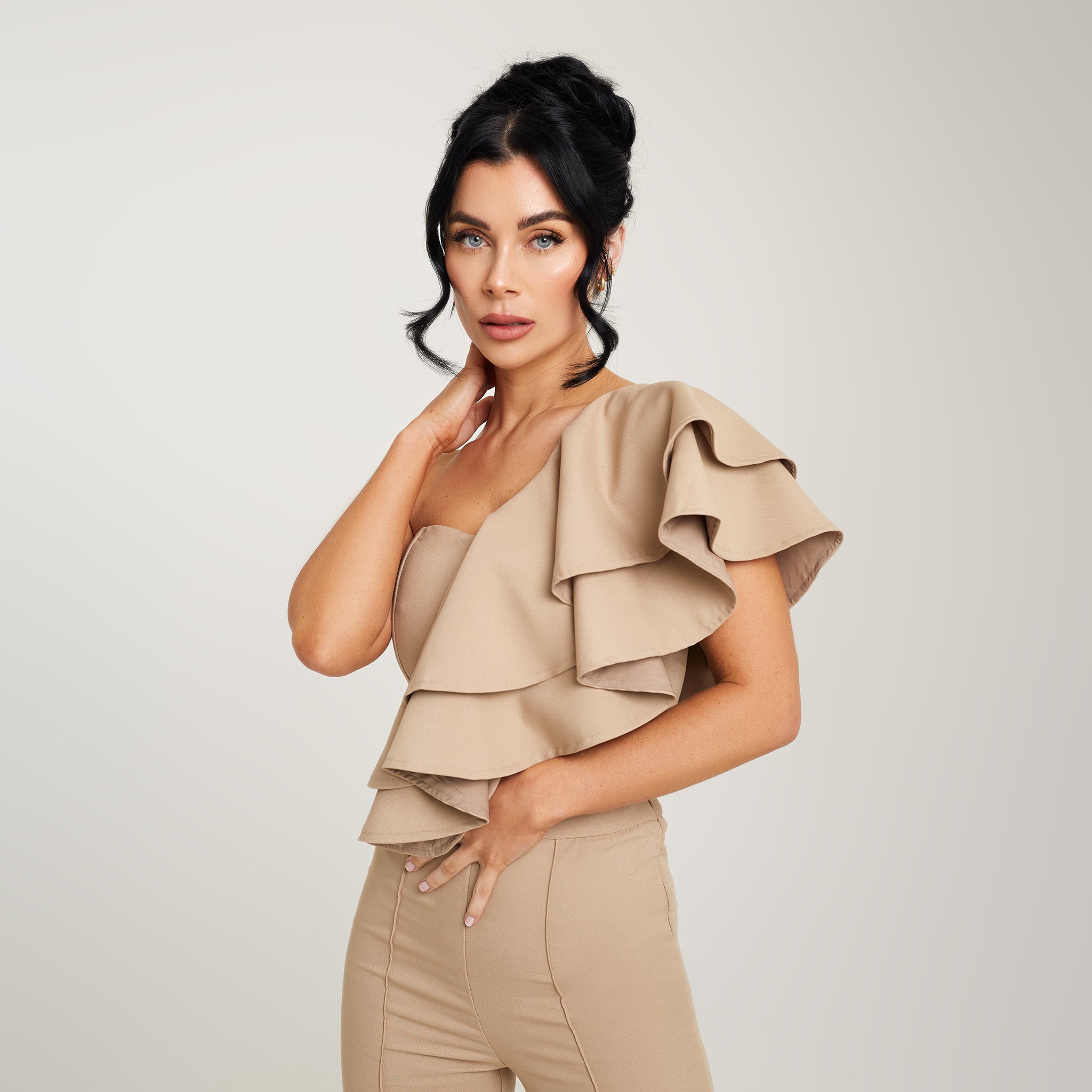 Cally Jane Beech, a woman with her hair styled up, models a beige one-shoulder ruffle cropped top. The top features a single shoulder strap, creating an asymmetrical neckline, while the ruffle detail delicately adorns the shoulder and extends down to the cropped hemline. The beige color adds a soft and neutral tone to the ensemble, offering versatility for various outfits.