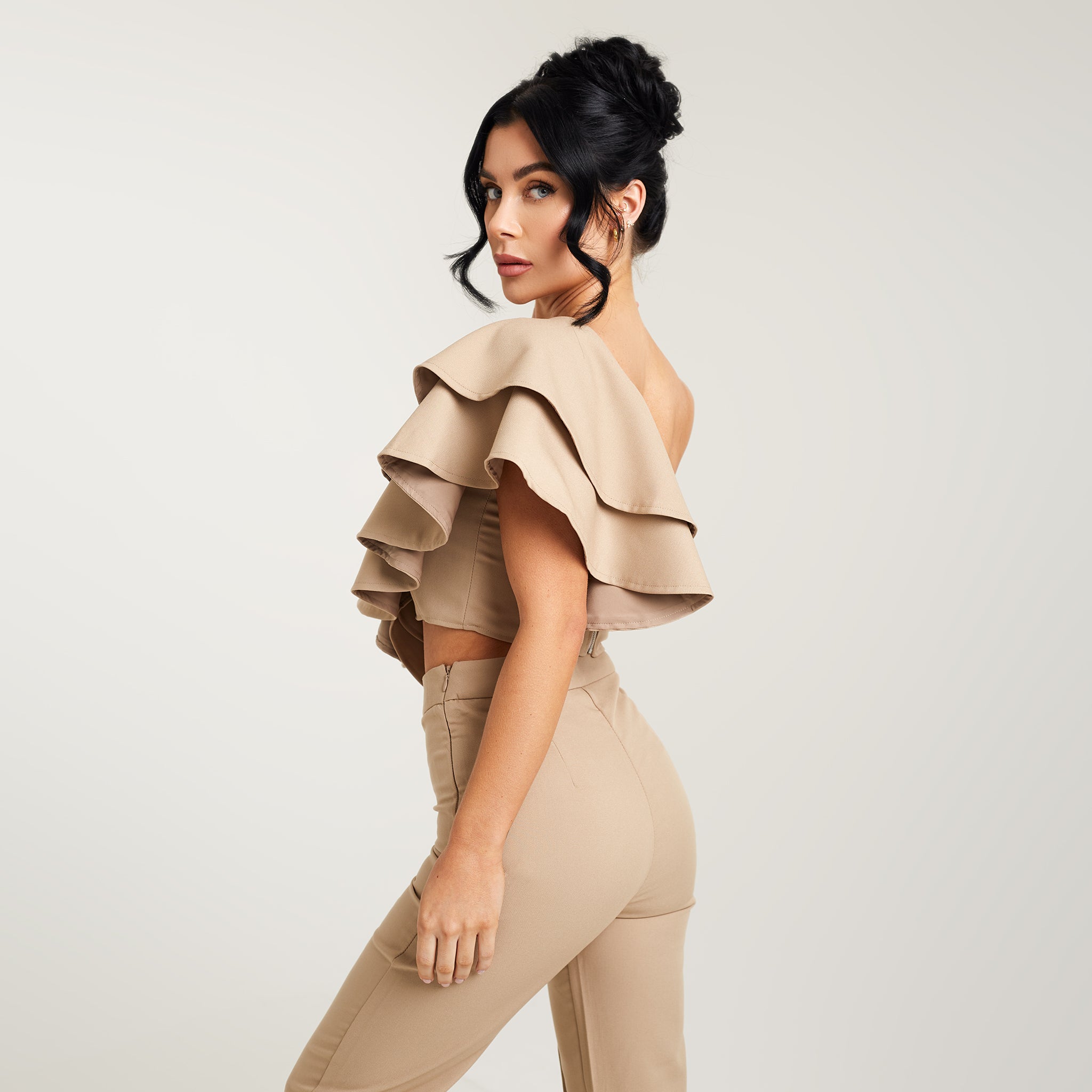 Cally Jane Beech, a woman with her hair styled up, models a beige one-shoulder ruffle cropped top. The top features a single shoulder strap, creating an asymmetrical neckline, while the ruffle detail delicately adorns the shoulder and extends down to the cropped hemline. The beige color adds a soft and neutral tone to the ensemble, offering versatility for various outfits.