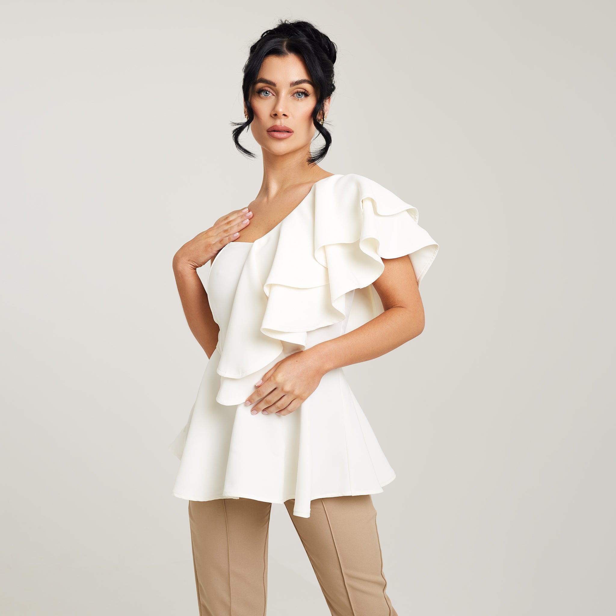 Cally Jane Beech, a woman with her hair elegantly styled up, models a cream-colored one-shoulder ruffle peplum top. The top features a single shoulder strap, showcasing an asymmetrical neckline, while the ruffle detail gracefully drapes down the bodice, creating a feminine and sophisticated look. The peplum waistline accentuates Cally Jane Beech's figure, adding a touch of flounce and elegance.