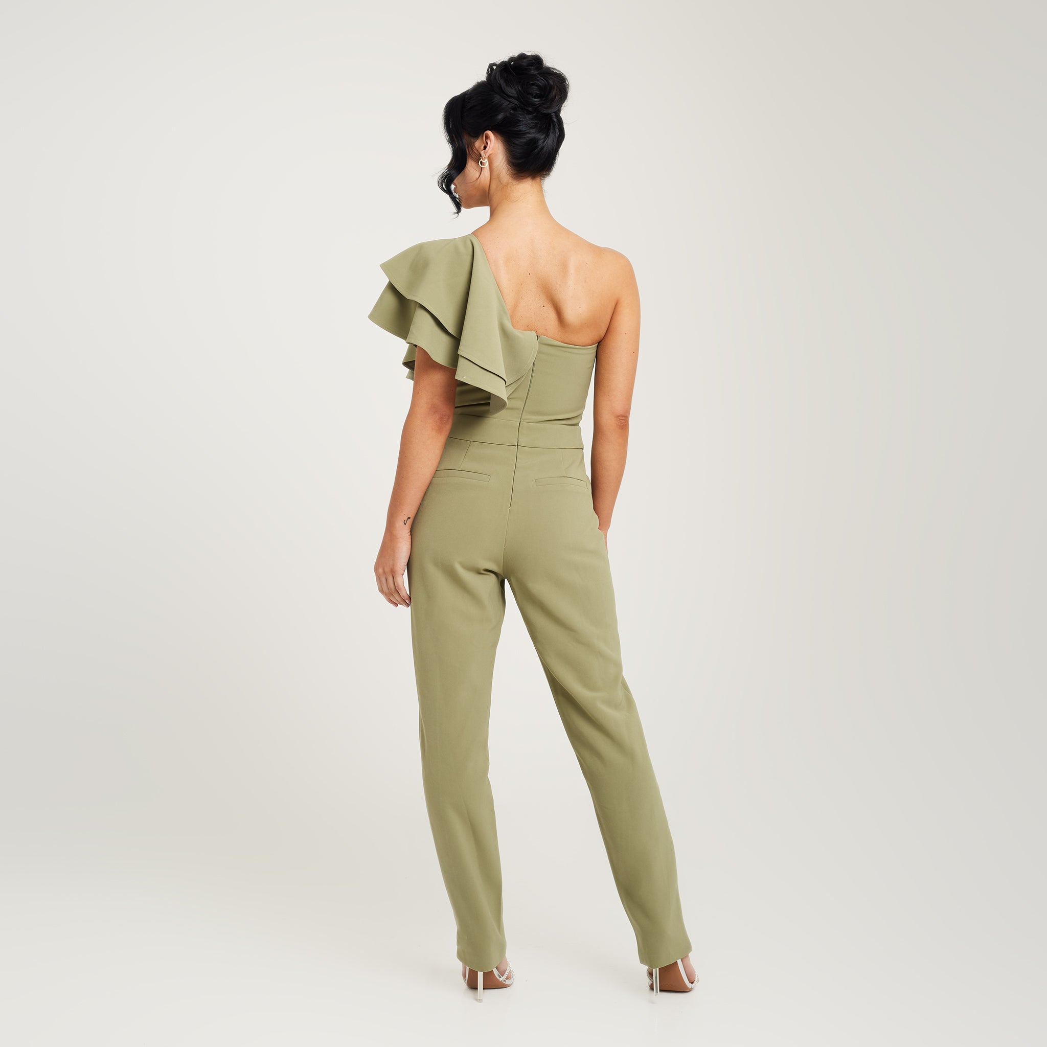 Cally Jane Beech, a woman with shoulder-length brunette hair, showcases a stylish sage green one-shoulder ruffle jumpsuit with cigarette trousers. The jumpsuit features a single shoulder strap, creating an asymmetrical neckline, while the ruffle detail cascades elegantly down the bodice, adding a feminine and playful touch. The jumpsuit's sleek cigarette trousers fit snugly and taper down to the ankles, creating a streamlined and polished look.