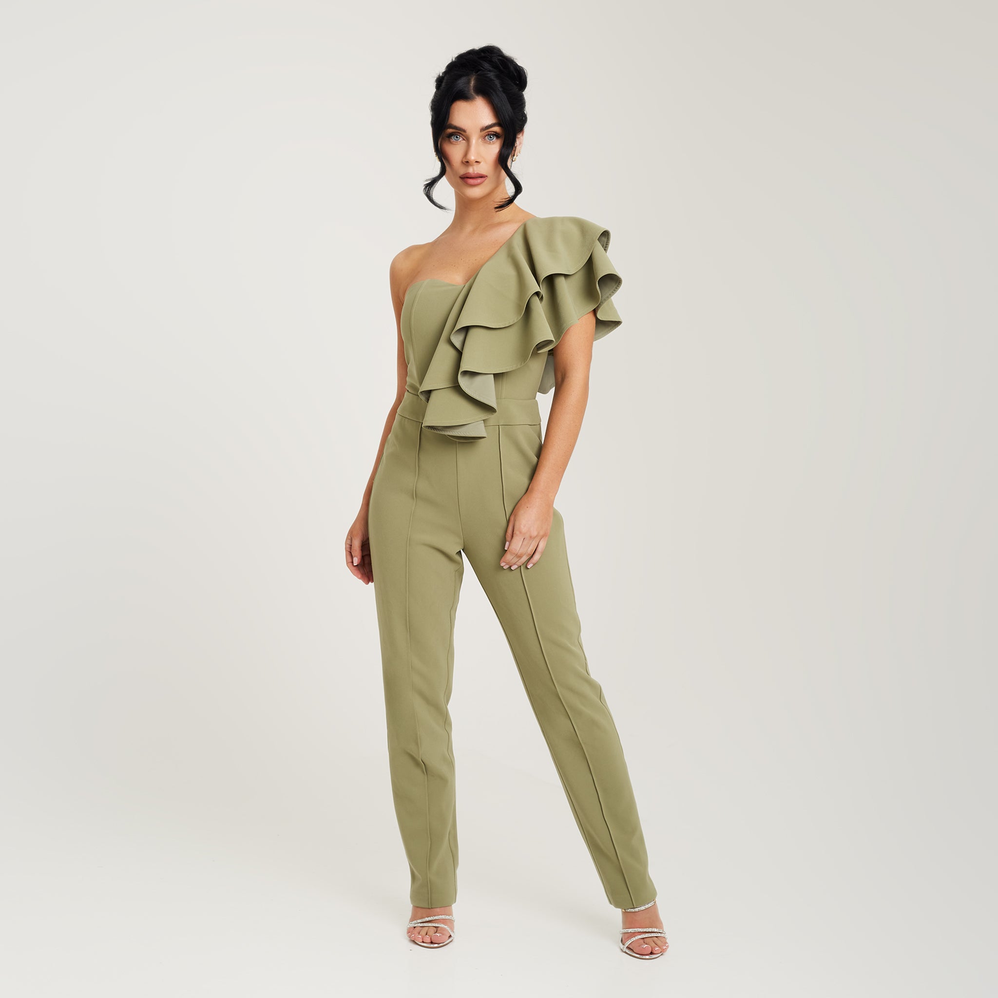 Cally Jane Beech, a woman with shoulder-length brunette hair, showcases a stylish sage green one-shoulder ruffle jumpsuit with cigarette trousers. The jumpsuit features a single shoulder strap, creating an asymmetrical neckline, while the ruffle detail cascades elegantly down the bodice, adding a feminine and playful touch. The jumpsuit's sleek cigarette trousers fit snugly and taper down to the ankles, creating a streamlined and polished look. Cally Jane Beech confidently wears the jumpsuit.